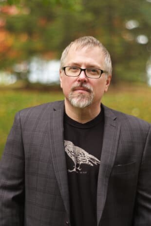 Jeff VanderMeer is a White man with hair and a beard that turn grey. He looks friendly yet stern. There's a bird on his t-shirt