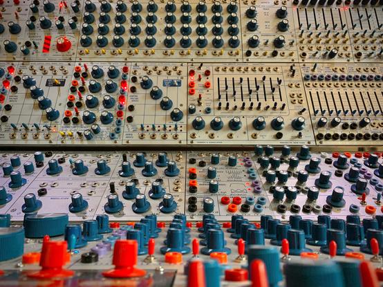 A large modular synthesizer (40U) Bucha clone system. There are many blue Rogan knobs with a few red knobs as accents. There are also a variety of plugs, metal sockets for audio and coloured plastic banana sockets for control signals. The module panels are grey with blue screen printed designs and text. There are a number of red LEDs illuminated throughout the panels.