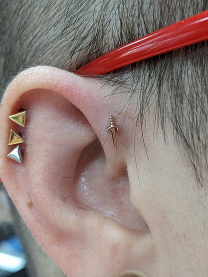A close of up a white human ear. The forward helix - the cartilage at the very front-facing-est part of the shell of the ear - is a little pinkish indicating it's freshly pierced, and sports a rose gold dagger. It joins a few other visible friends - a trio of polished metal triangles in the helix and just the edge of a stretched lobe are visible.