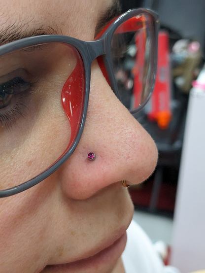 A white human's face in semi-profile. The human is wearing glasses and sporting a brand new nostril piercing - a stud with a tiny round garnet gem set in silver. The new stud joins a rose gold septum ring already deployed. The human also wears slightly oversized glasses and a thoughtful expression.