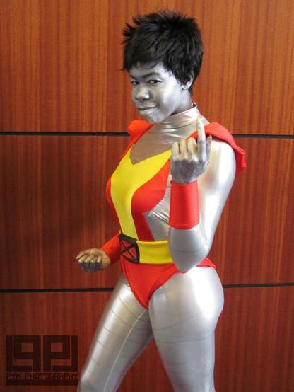 A Black woman with silver body paint, silver bodysuit with segmented arms, legs, and chest, wearing a red and yellow leotard with a yellow belt and a black and red X-men buckle on the front, standing in front of a wooden paneled wall. The watermark on the bottom left says Pym Photography