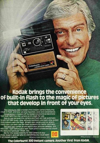 Dick in a green turtleneck smiles with a flashing kodak camera.