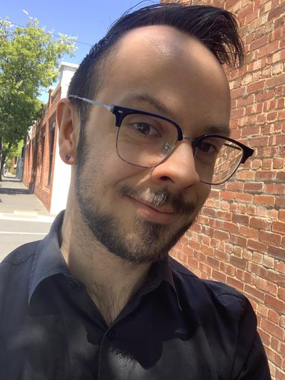 Photo of a man’s face. He’s showing off a metallic purple earring.