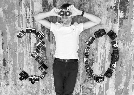 In the photo, a man is lying on the ground. He is holding two lenses and looking through them. To the left and right are cameras placed in the shape of the numbers "3" and "0".