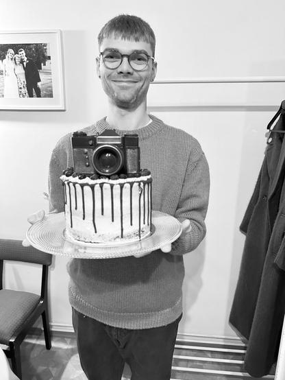 In the photo, a man holding a cake with a Zenith chocolate camera. He is wearing chunky winter sweater and glasses.