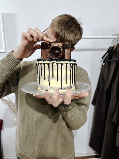 In the photo, a man posing for a photo with a cake. He pretends to take the picture with a chocolate camera.