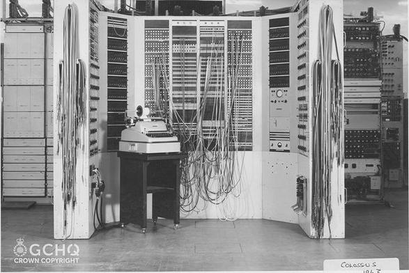 A photo of one of the earliest digital computers from the Colossus line. In the foreground is a curved array of panels with many lights and patch cables along with a text terminal.