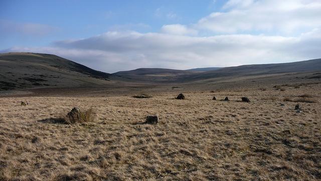 A ring of small upright stones on a flat moorland plateau of scrubby grass. Hills rise gently on both sides and in the background. A further circle lies just beyond, but is difficult to see as the stones are also small. The sky is blue, with some high cloud. It's very cold.