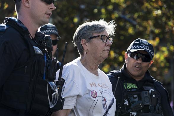 A woman with grey hair and wearing a white protest t shirt is arrested by several police officers.