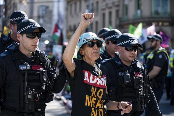 An older woman in a blue hat, sunglasses and black tshirt which says "Adani, no means no" raises her fist while being arrested by five police officers.