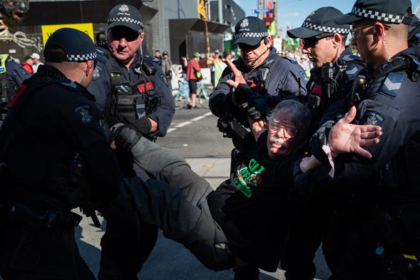 A grey-haired man with glasses being carried from a protest by five police officers on a sunny day.