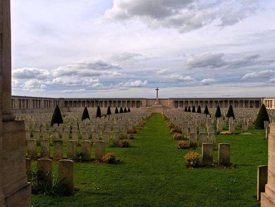 View from the entrance. The site consists of a colonnade of wall panels forming three sides of the perimeter of a cemetery, and incorporating a Cross of Sacrifice. The names of the missing are inscribed on the panels, arranged by regiment or other unit. Along the sides and the back, stone tablets are fixed in the stone rubble walls bearing the names of the dead grouped under their Regiments. On a lawn are plenty of gravestones plus some trees. There is a gloomy, desolate silence in this place.