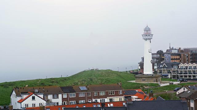Photograph of a lighthouse on the right side of the photo, surrounded by low houses on three sides. On the left, gray sky and sea which are so much the same color you can't distinguish them.