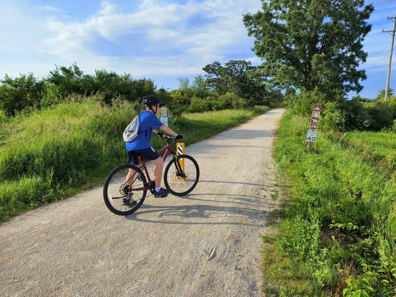 The start of the 51.3 mile ride. Cyclist in a blue shirt on a red bike. Bike path made of crushed limestone and recedes off into the distance. The path is lined with native plants and trees.