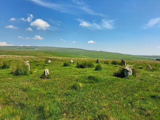 A ring of 9 granite standing stones standing on grassy moorland (more smaller stones are hidden in the grass). The ring surrounds the remains of a Bronze Age cairn, now a very low mound. Reedy grass intrudes around the stones. A high green ridge rises beyond the stones on the skyline (Gutter Tor). The sky is deep blue with some wisps of white cloud. It's too hot to think. This is Ringmoor Down cairn circle.