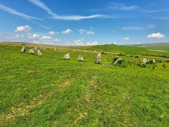 A wide, spacious open ring of granite standing stones on gently sloping grassy moorland. This is Brisworthy circle. The ground slopes gently down to the right and the circle is built on the slope. Beyond a rocky tor rises (Legis Tor), an obvious focal point from the circle. Beyond that, on the far right skyline higher ground rises. The sky is very blue with some scattered cloud. It's so hot my brain has melted.