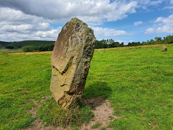 A large slab of reddish sandstone stands in a cropped grass field. It leans to the right. A further, much smaller stone stands  in the background to the right. The scene is sunny but the cloud cover is thickening on the left of the blue sky.