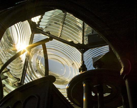 Image of the inside of a fresnel lens in a lighthouse. The interior metal framing is silhouetted against the concentric circular lenses, and the Sun is shining through one of them.