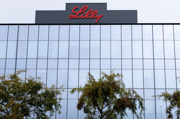 Photo of the HQ building of Eli Lilly in Boston, MA. The top signage has the familiar red cursive rendering of the name "Lilly" against a black background. Below it are mirrored office windows and a canopy of green trees.