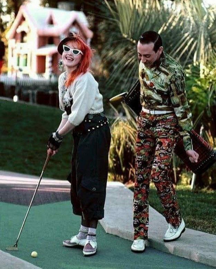 Cyndi Lauper is about to swing at a golf ball while PeeWee Herman stands behind her