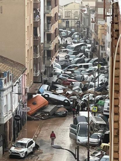 A photograph of a street in Spain. On either side of the street are houses and buildings. In the street are piles of washed up cars and vans, with a few people in the street, looking at the mess.
