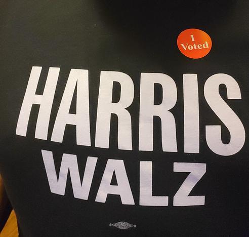Close up selfie of a Harris Walz t-shirt with a red dot I Voted sticker on it. A Union Made logo is visible on the shirt as well.