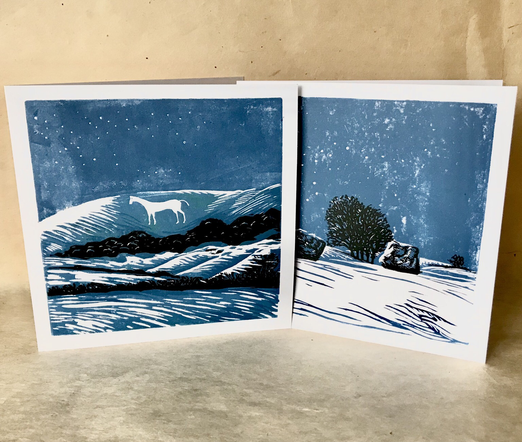 Photo of two handmade lino prints showing night time scenes of the Westbury white horse in a snowy blue and white landscape with dark trees and shrubs, and two standing stones from the Avebury circle backed by tue henge bank and tall trees. In both the sky and shadows are in blue and flakes of snow are flowing across the sky