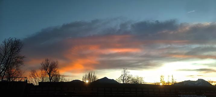 A breathtaking sunset scene featuring a mountain range silhouetted against a vibrant sky. Warm tones of orange and pink streak across the clouds, blending with the deepening blue of the twilight. Bare trees frame the foreground, their branches stretching upward, adding a dramatic contrast to the glowing horizon. The last rays of sunlight peek between the mountains, creating a serene and magical atmosphere.