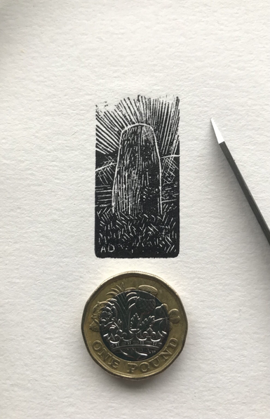 Top-down photo of a tiny wood engraved,black ink print of a standing stone in a landscape with dynamic lines denoting the sky. Upon the print is  a £1 coin and a cutting tool, for scale. the printed area is 37mm tall by 20mm wide. The initials AD are in the bottom left of the design