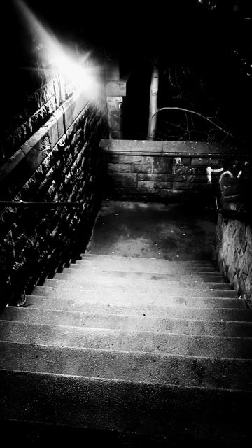 Looking down a flight of stone steps descending the side of a bridge, at night, one streetlight overhead creating a pool of light surrounded by shadows