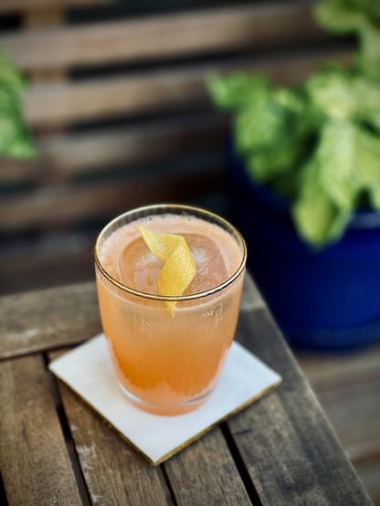 pinkish orange cocktail with lemon twist garnish sitting on a white coaster on a wooden table with plants in the background