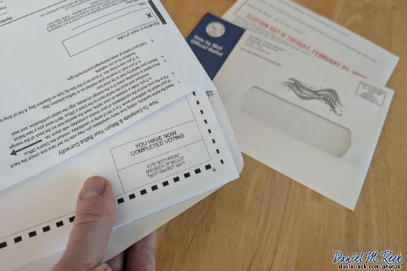 A voter holds a mail-in ballot, mostly inserted into the envelope. The corner of the ballot reads, "You have now completed voting." Photo by Daniel M. Reck, danielreck.com/photos