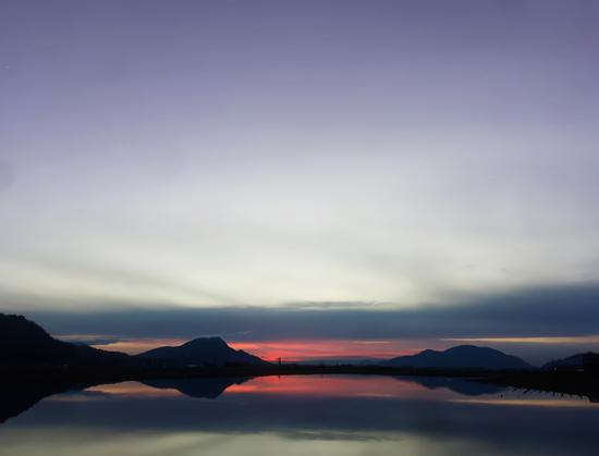 A serene sunset scene featuring a tranquil body of water reflecting the gradient sky above. The sky transitions from soft purples and blues at the top to warm oranges and pinks near the horizon. Silhouetted mountains frame the reflection, creating a harmonious balance between the natural landscape and its mirrored image.