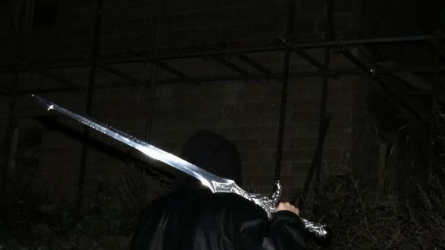 A very dark image where you can guess someone from the back with a black cape. They are holding a very big and bright silver sword.