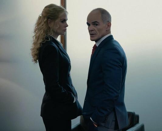 Nicole Kidman and Michael Kelly in business attire have a conversation in an office hallway.