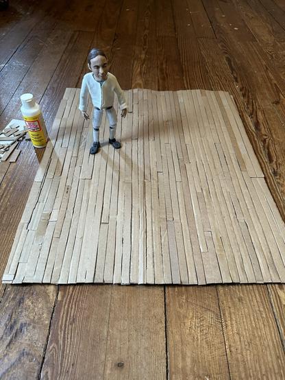 a stopmotion puppet stands on a miniature wood floor made of cardboard, mod podge, acrylic, and watercolor pencil