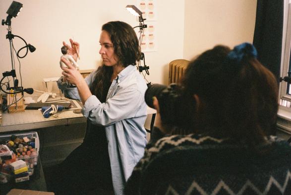 a female puppet maker works on a claymation puppet and is photographed by another woman in the foreground