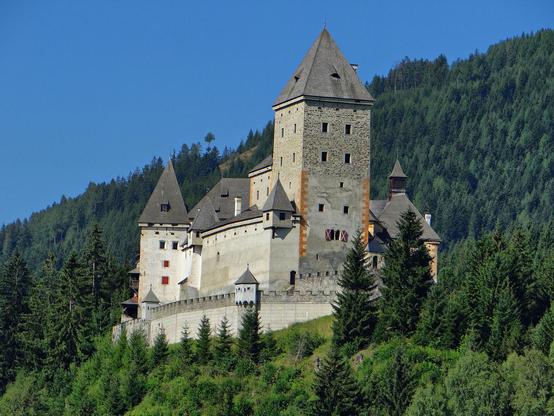 a  photo of a castle built along a river with a mountain behind it, it has large square towers with big pointed tops, the body of it is tan, the roofs grey-black