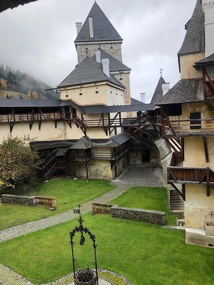 a photo of a courtyard area in the castle, it is very medieval and cool looking, the walls  a light napkes yellow, there s a lawn , and a well for water