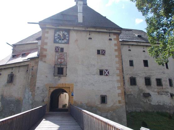 a photo walking into the castle on a wooden bridge, there is a clock on the outside of the wall, there are small windows in the thick walls