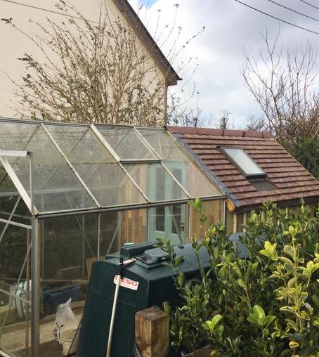 A picture of a greenhouse attached to a shed.

The greenhouse is a standard 8 feet by 6 feet aluminium-framed model, but a purpose-built shed has been built in line with it so that the roofs line up. The roof of the shed is covered with brown slates and a copper ridge, and there is a skylight in the middle. Internally, the shed and greenhouse are separated by a duck egg blue door. The sides of the shed are wooden.

The photo is taken diagonally from one side. In the foreground is a green oil tank, and in the background the white side elevation of next door’s house.