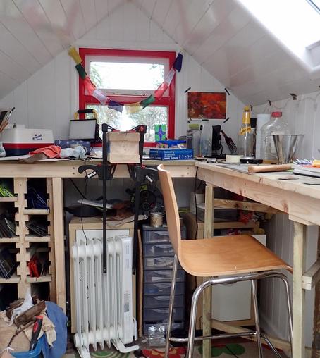 The inside of the shed described in the first photo. 

The shed is a workshop, with a workbench running transversely along the back and down one side under the skylight. Underneath, on the left, is a rack system containing pieces of coloured glass. In the middle of the bench, two clamps are attached to a block of wood. On the back wall is a window, with a red frame.

A chair is parked in the middle. Mostly, it’s a good, chaotic mess.