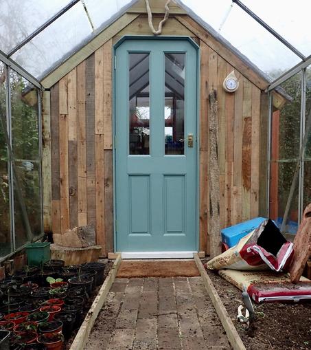 The inside of the shed greenhouse, taken from the greenhouse section of the entrance to the shed. It’s a wooden wall, with patchwork panels oriented vertically and in the middle, a duck egg blue door, half-glazed.

A slabbed path runs up the middle, with growing beds of soil on either side. Plastic pots and other detritus are scattered around.