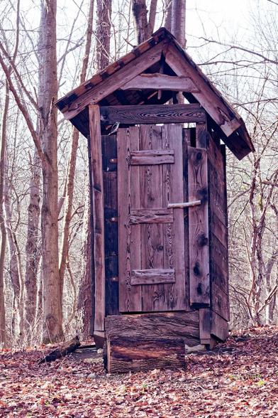A tall wood outhouse made of charred wood with a lopsided roof.