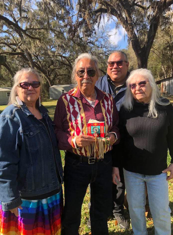 A photograph of Leonard Peltier, who now has white hair, standing with friends and relatives. He is wearing a traditional shirt and black jeans, and is smiling.
[Photo credit Gina Buenrostro]
