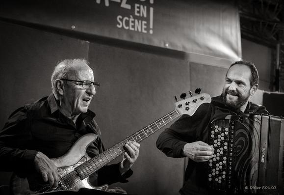De 3/4 en plan américain, Christian à gauche joue de sa basse en regardant droit devant lui, tandis que Frédéric le regarde en souriant et en jouant de son accordéon(photo en noir et blanc)