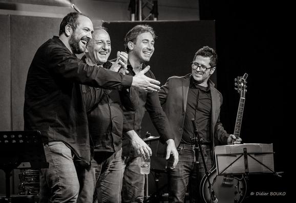 De 3/4 en plan américain, tous les musiciens sourient, Frédéric son bras droit tendu vers le public, Christian ses mains jointes, Cédric ses deux mains vers le bas et Filipe regarde vers les autres musiciens sa guitare dans sa main gauche et sa main droite dans le dos de Cédric à sa droite (photo en noir et blanc)