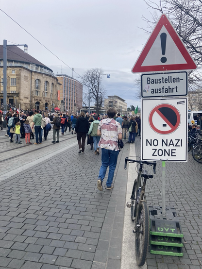 Auf einer Straße mit vielen Menschen steht ein Schild „No Nazi Zone“, daneben lehnt ein grünes Rad