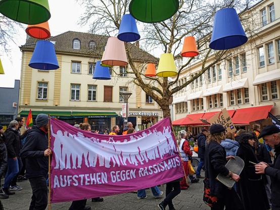 Demozug unter bunten Lampenschirm. Im Vordergrund ein Antirassismus Plakat.