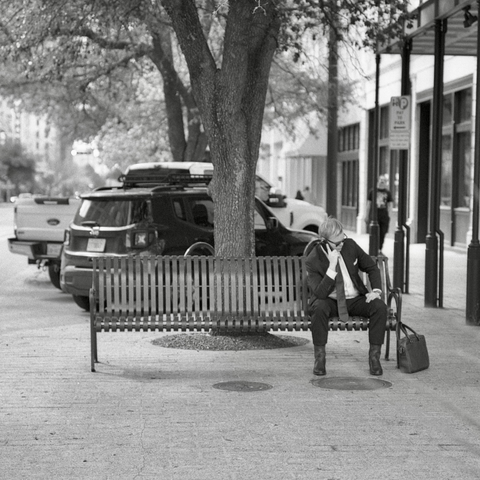 A black and white film photo. A man in a suit, seated on the far side of a city bench, takes a phone call in downtown Austin, Texas. Tuesday, February 4, 2025.